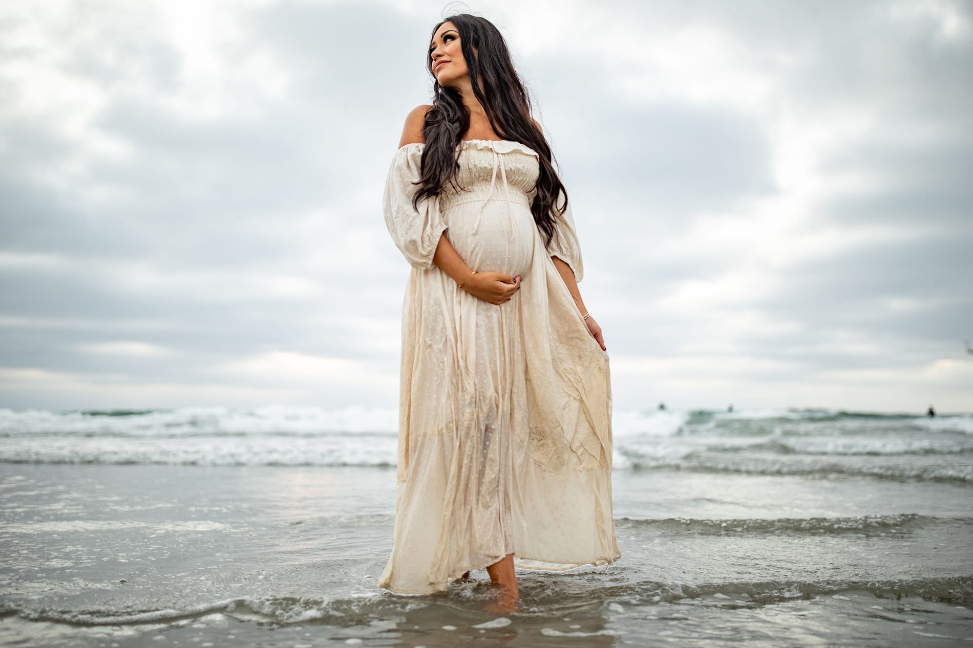 Maternity Photographer, an expectant mother stands in shallow beach waves holding her belly, she wears a white dress