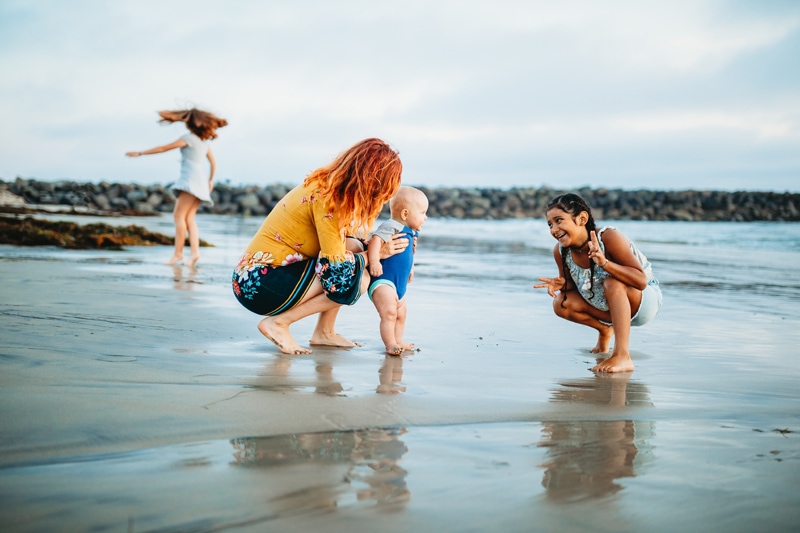 Family Photographer, a mother holds her toddler as her older daughters play beside her on the beach sand