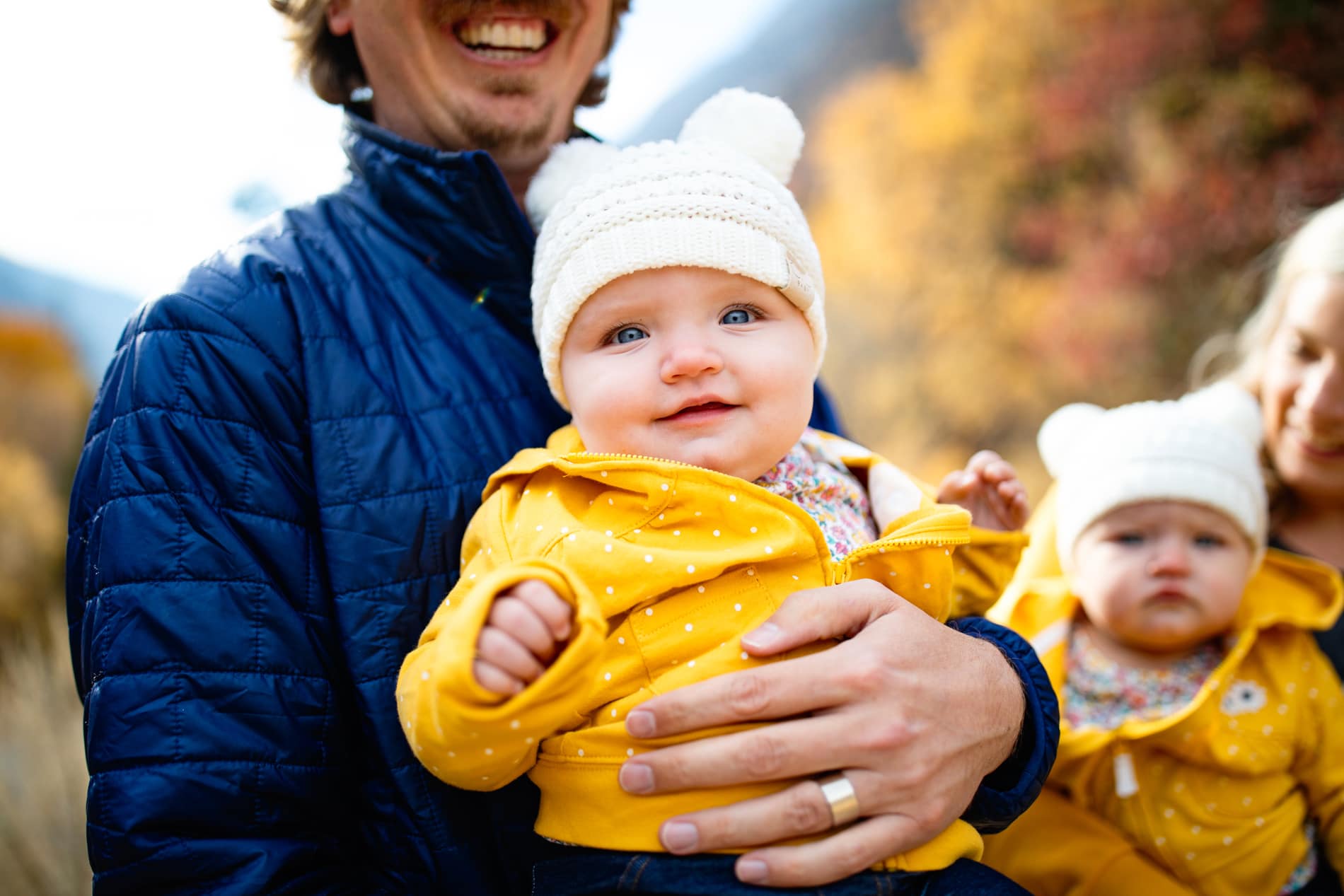 Family photographer, a father holds his smiling baby. Baby wears a yellow coat and knit beanie.