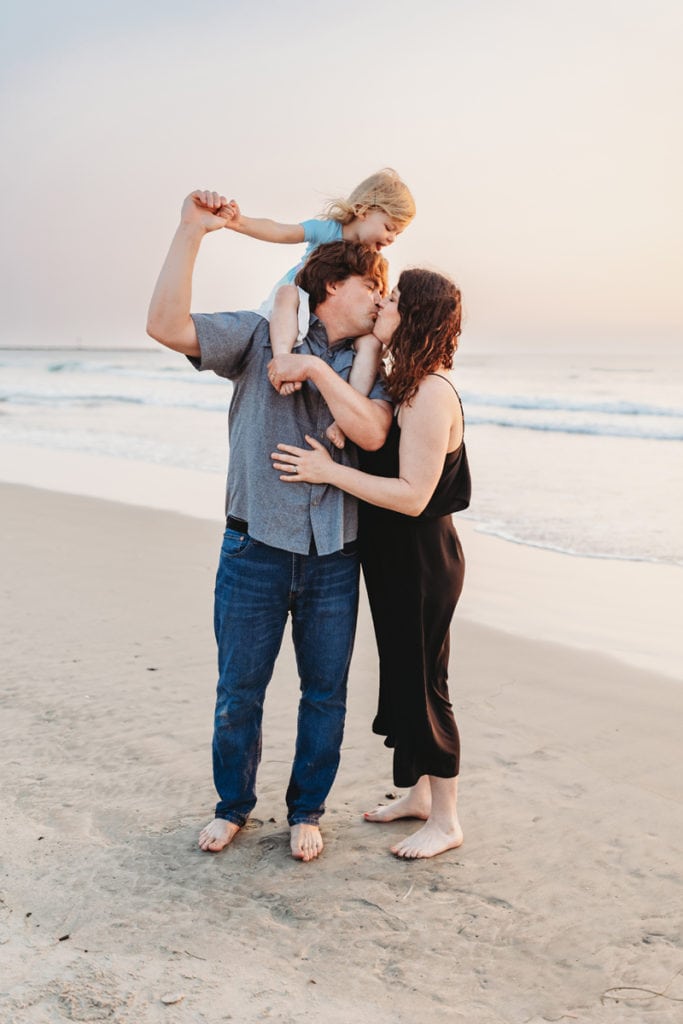 Family Photographer, a husband and wife kiss, a smiling toddler sits on her dad's shoulders. They are at the beach.